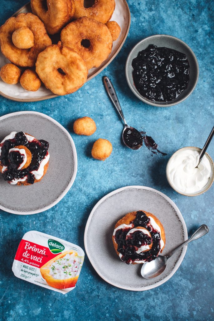 Papanași prăjiți aurii, serviți cu topping de smântână și dulceață de fructe.