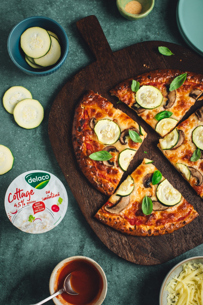 Felie de pizza cu blat de brânză cottage, mozzarella, ciuperci și dovlecei, alături de Brânza Cottage Delaco.